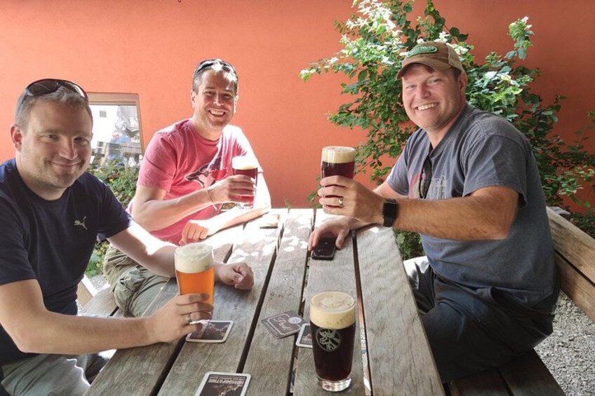 Three people sitting at a wooden table outdoors, holding glasses of beer. 4