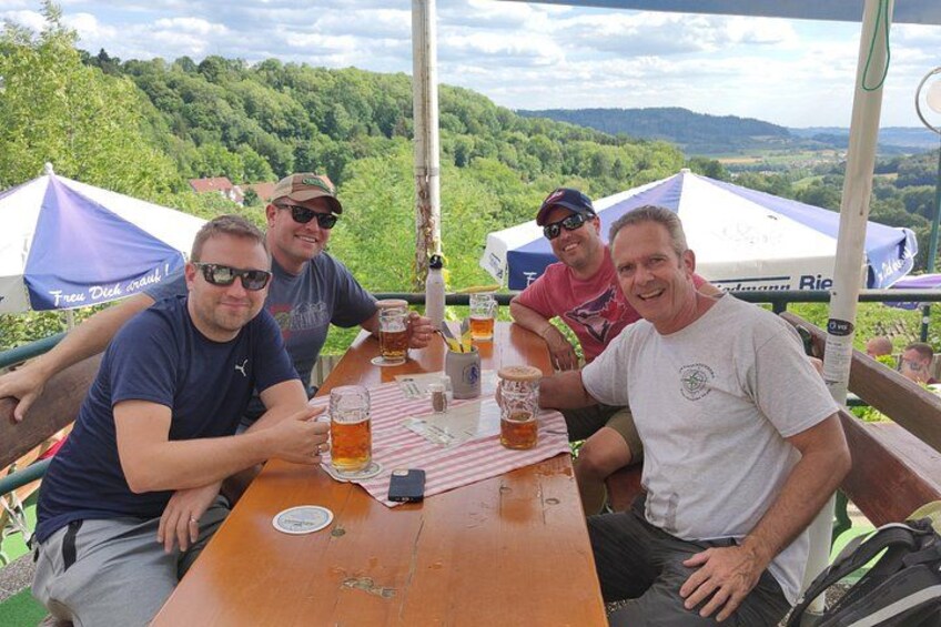 Four people sitting at a wooden table outdoors with a scenic view of hills and trees in the background. 2