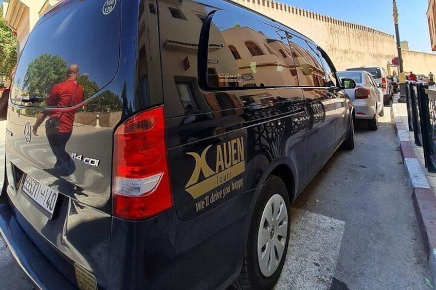 Un fourgon noir est garé dans une rue dont la chaussée est réfléchissante. On aperçoit un homme portant un t-shirt rouge, et un bâtiment en arrière-plan. 3
