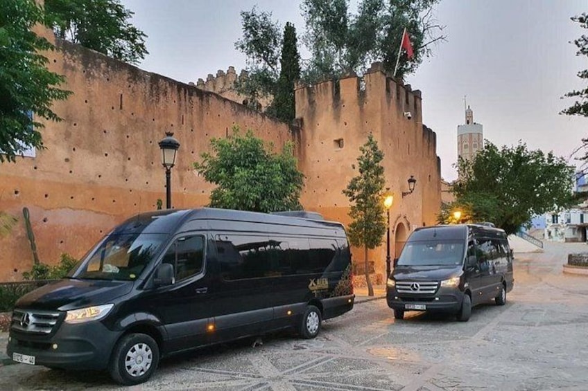 Twee zwarte bestelwagens geparkeerd voor een historische stenen muur, met een lantaarnpaal en bomen. 3