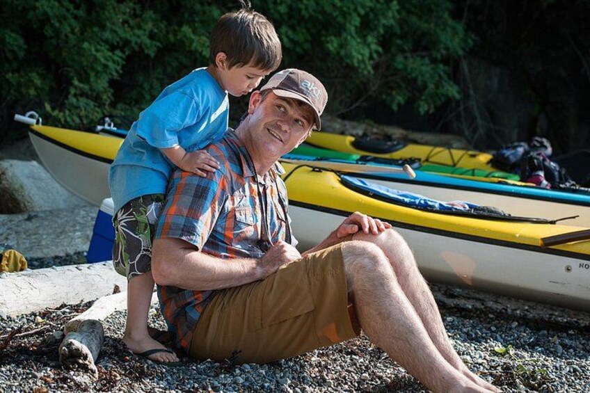 A man sitting on a beach with a child on his shoulders, kayaks in the background. 4