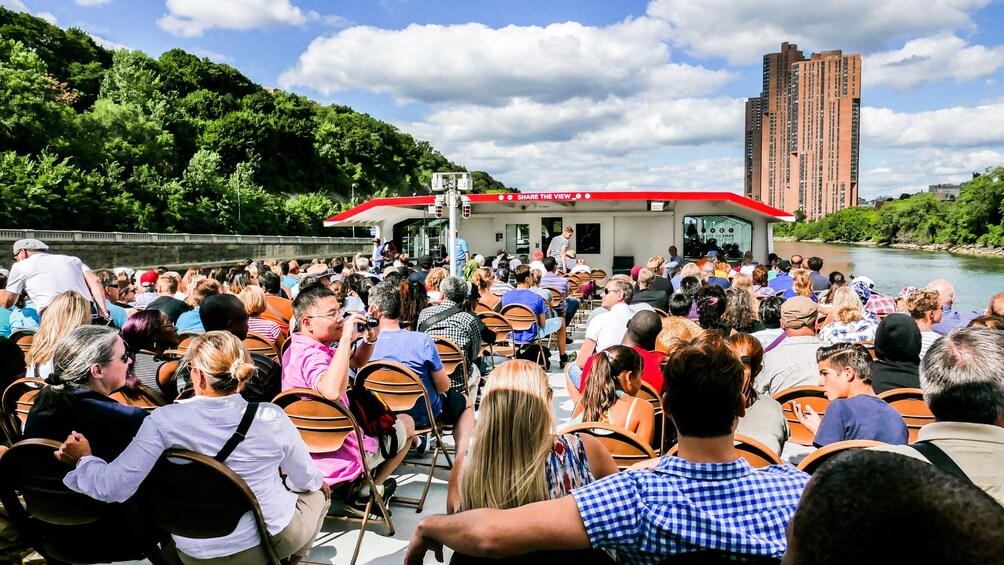 A crowd gathered on a boat, facing a stage with a red roof, overlooking a river with a cityscape in the background. 3