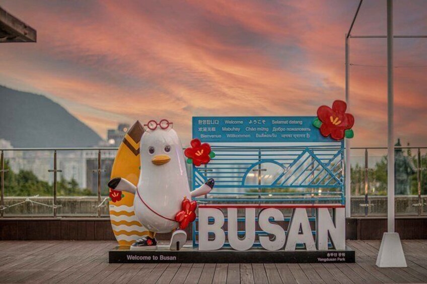 A bus stop sign with 'BUSAN' and 'Welcome to Busan' text, a decorative bird figure, and a cityscape with mountains in the background. 5
