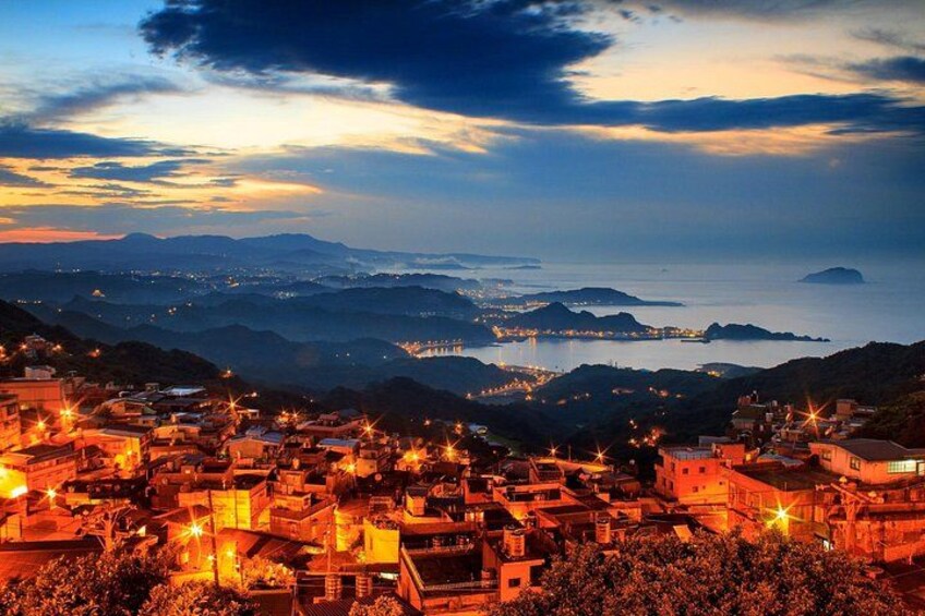 Un pueblo costero al anochecer con edificios iluminados y vistas al mar y montañas distantes. 5