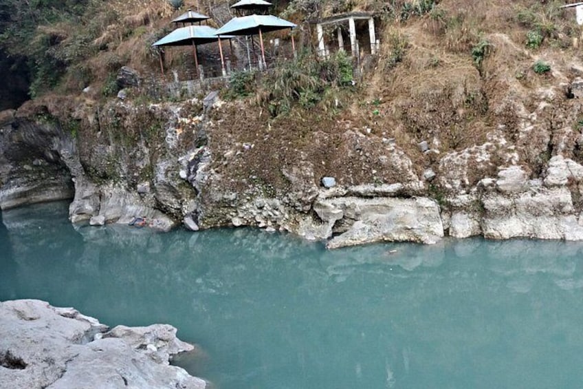 A rocky cliff with a turquoise river below, a small structure with a roof, and some vegetation. 5