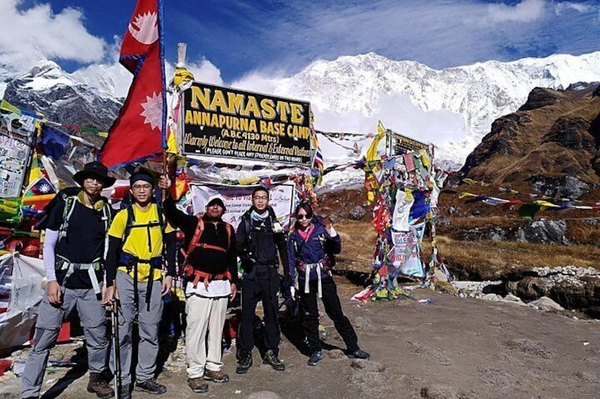 Un groupe de personnes se trouve au camp de base, au pied de la montagne, devant une pancarte sur laquelle on peut lire « NAMASTE, CAMP DE BASE DE L’ANNAPURNANA », et un drapeau rouge flotte. 3