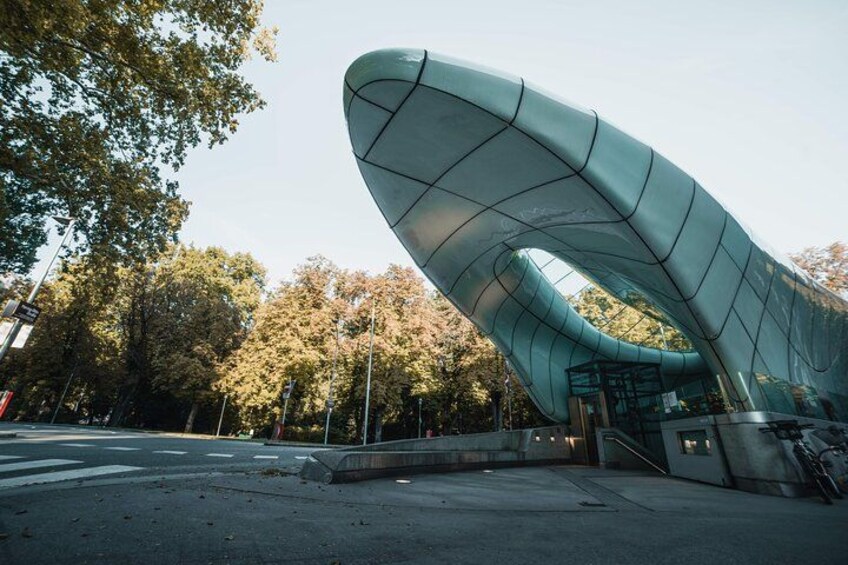 A modern architectural structure with a curved roof, surrounded by trees and a paved area. 2