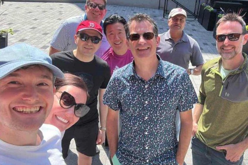 A group of people standing outdoors, some wearing casual clothing and hats, with a paved area and plants in the background. 3