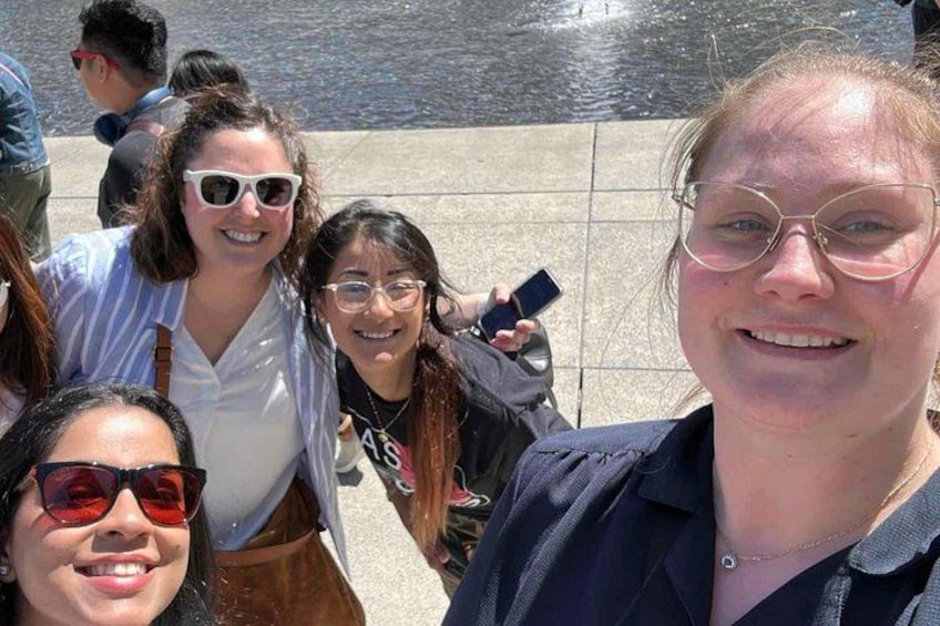A group of people standing outdoors, possibly at a public event or gathering, with a body of water and tiled pavement in the background. 2