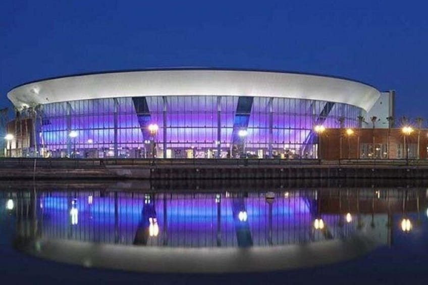 A modern stadium with a curved roof, illuminated by purple lights, reflecting on the water in front. 4