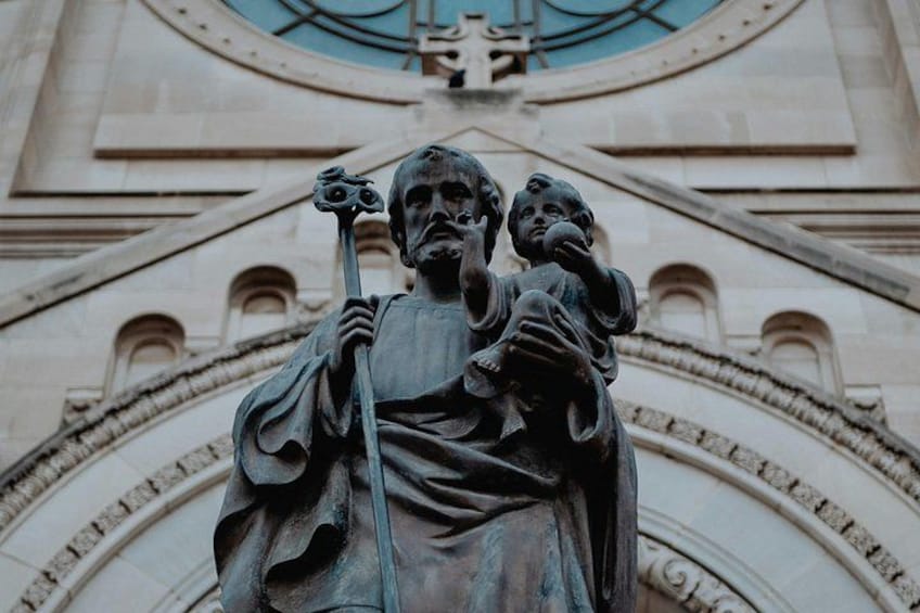 A statue of a figure holding a child and a staff, set against a building with arched windows and decorative stonework. 1