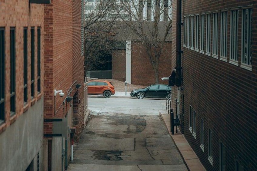 A narrow alleyway with brick buildings, a parked orange car, and a black car. 2