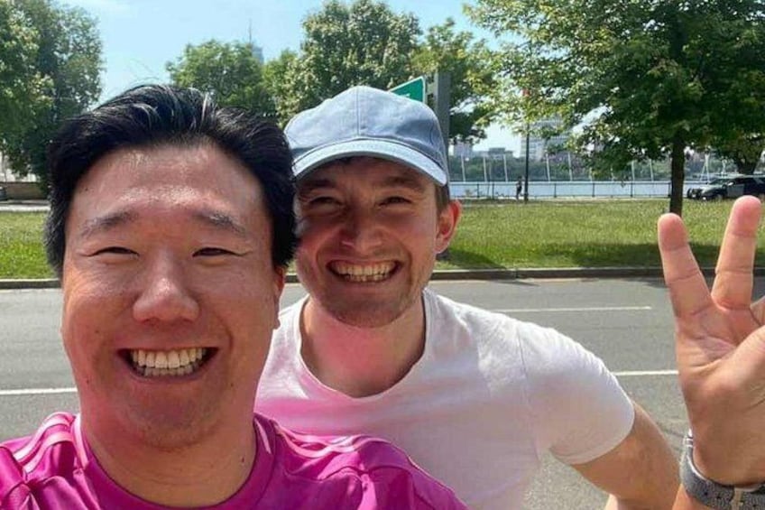 Two people outdoors, one in a pink shirt and the other in a white shirt and cap, with a road and greenery in the background. 2