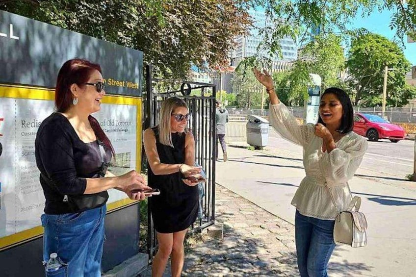 Three women standing outdoors near a bus stop with a signboard. 2