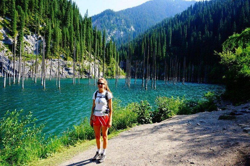 A person standing on a dirt path beside a lake surrounded by trees. 4