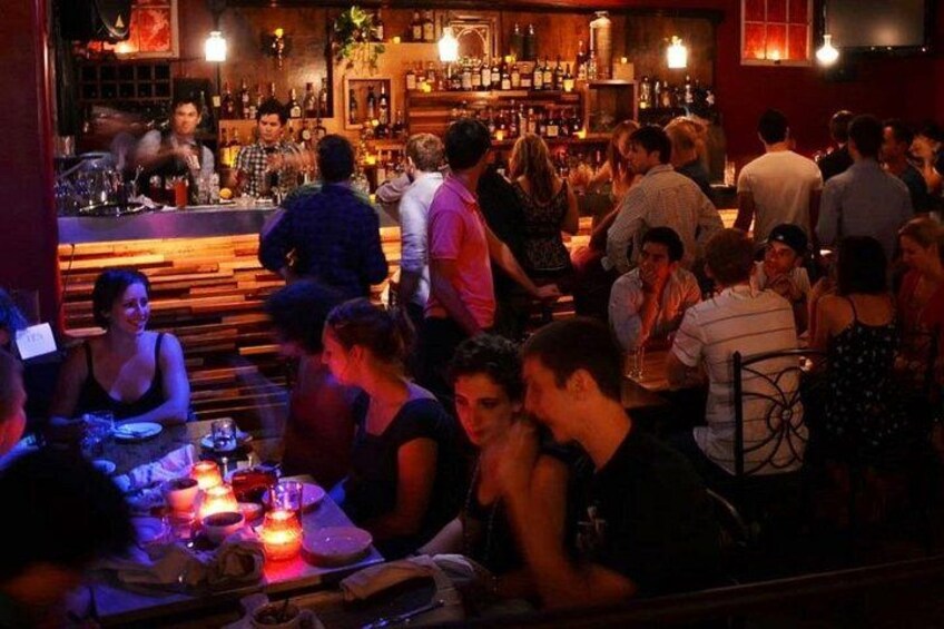 Un bar concurrido, con gente sentada y de pie, un camarero detrás de la barra y diversas botellas y vasos en estantes. 3