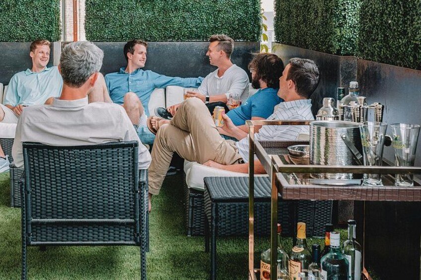 Un grupo de personas sentadas en muebles de exterior, una zona de bar con botellas y vasos, y un seto verde al fondo. 2