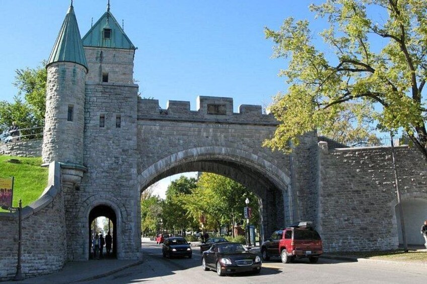 A stone archway with a tower, vehicles passing through, and a tree in the foreground. 4