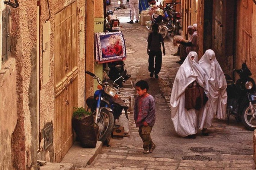 A narrow street with people walking, a child, and parked motorcycles. 1