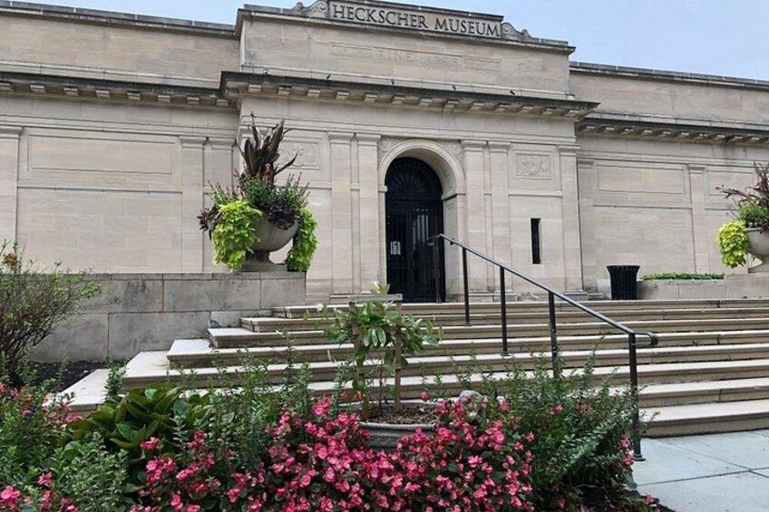 The exterior of the Heckscher Museum with steps leading up to the entrance, surrounded by greenery and flowers. 4
