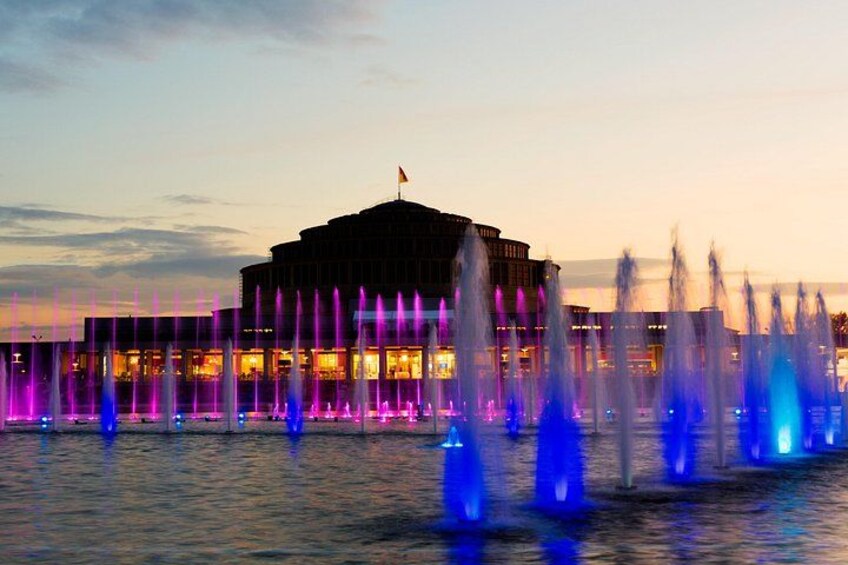 A building with a flag on top, surrounded by water fountains with colorful lights. 2