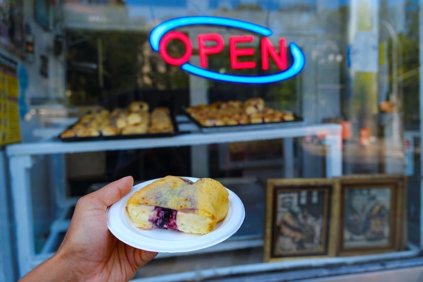 Una mano che tiene un pasticcino con ripieno visibile, davanti a una vetrina con un neon 'OPEN'. 1