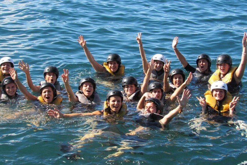 Eine Gruppe von Menschen im Wasser, die Rettungswesten und Helme tragen, wobei einige die Hände heben. 3