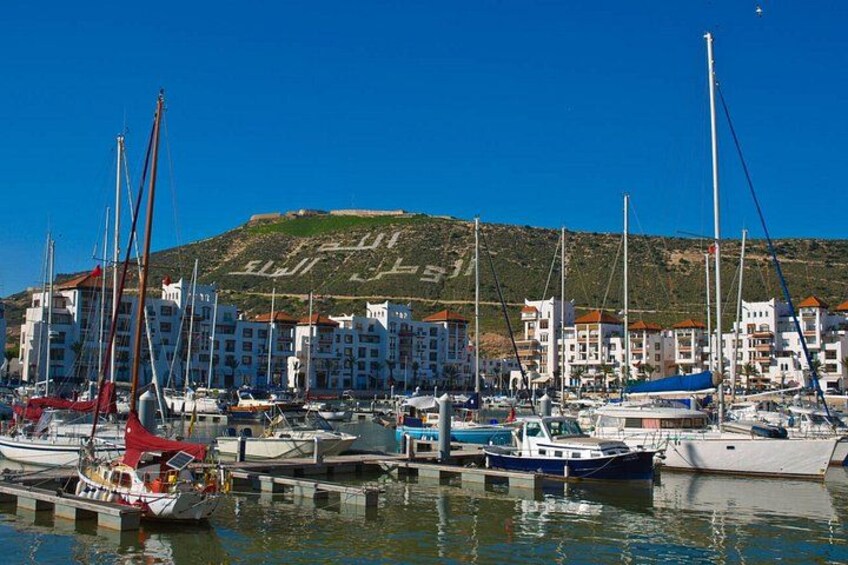 Un puerto deportivo con barcos amarrados, una colina con edificios al fondo y cielos azules despejados. 2