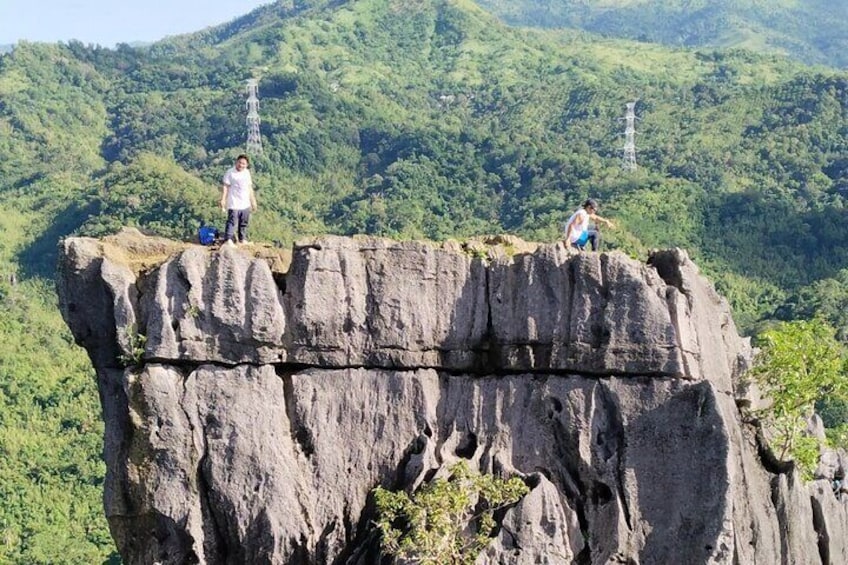 Two people standing on a rocky cliff with a forested mountain in the background. 1