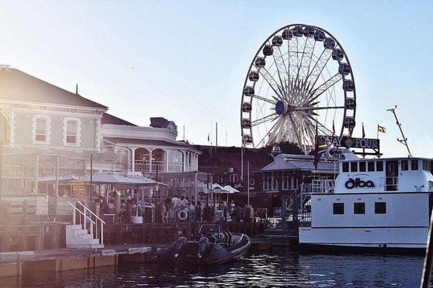 A waterfront with a Ferris wheel, a boat docked at the pier, and buildings with outdoor seating. 4