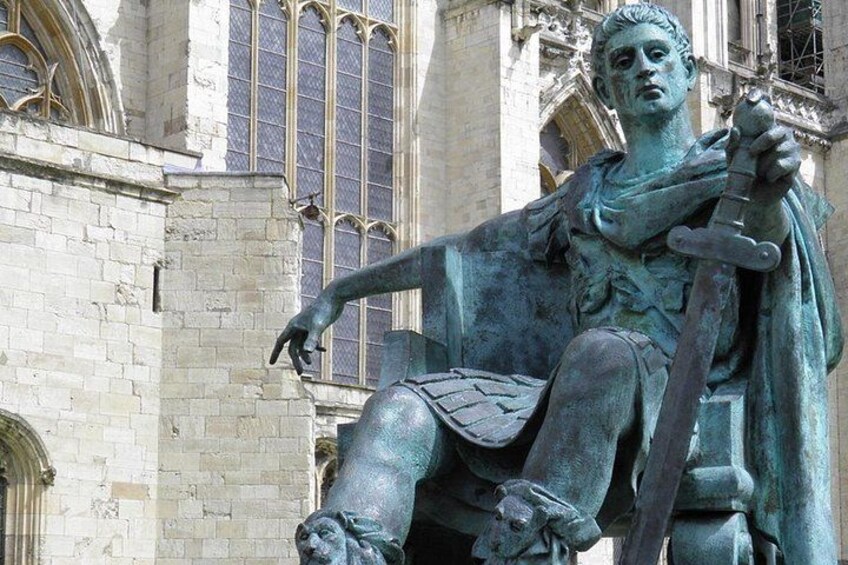Een bronzen beeld van een historische figuur, zittend op een stenen sokkel, met gotische architectuur op de achtergrond. 1