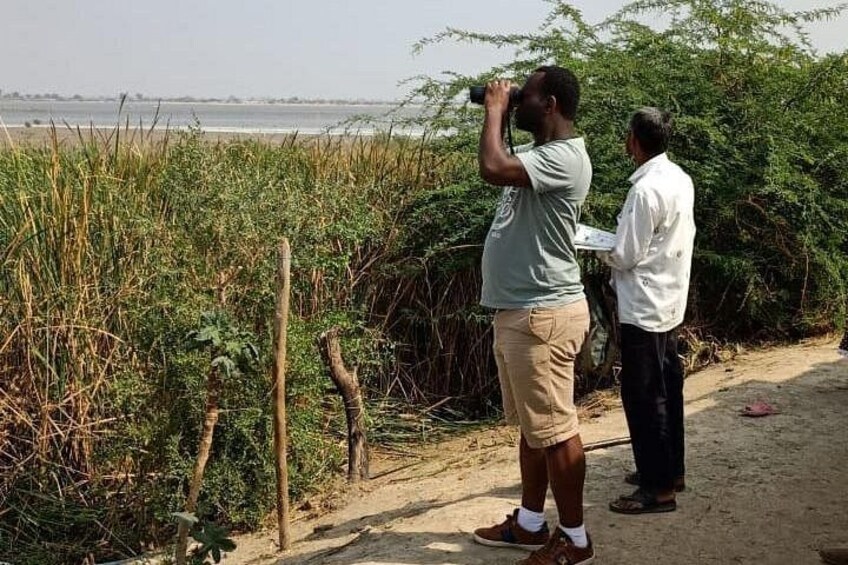 Two people standing by a riverbank, one using binoculars. 3