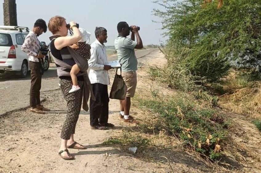 A group of people standing on a dirt road, some holding binoculars, with a vehicle parked on the side. 4