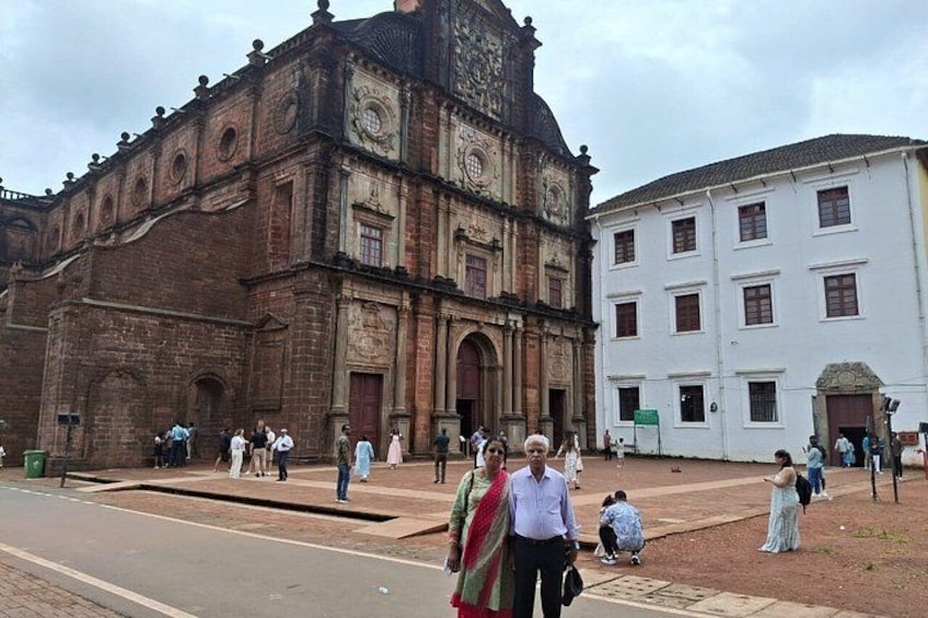 A historic building with intricate architecture, a paved area with people, and a white building in the background. 2