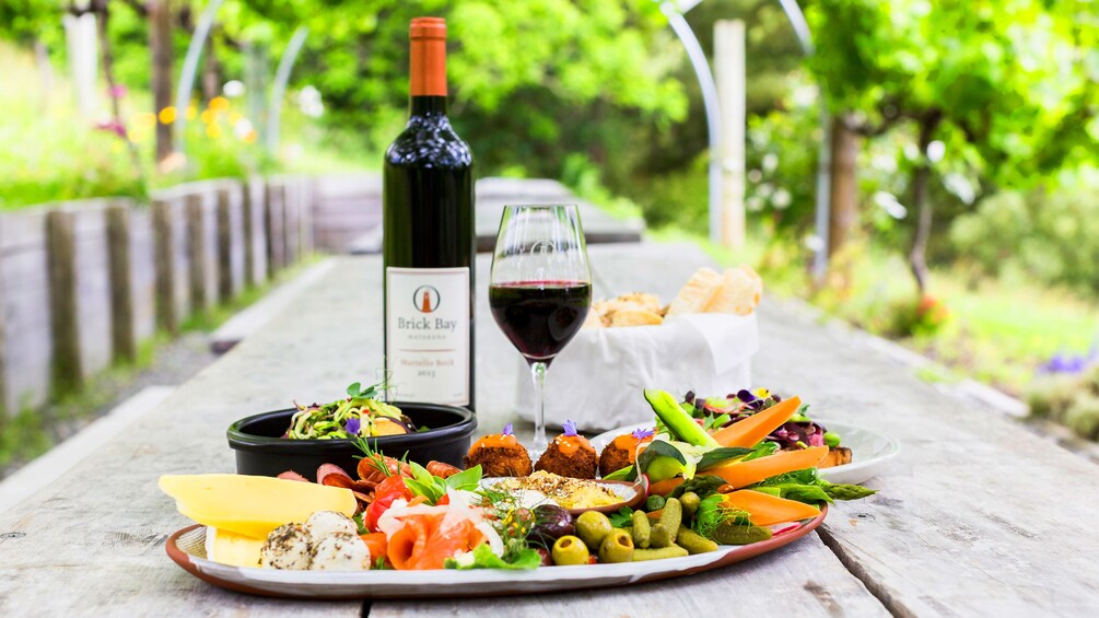 A picnic table with a bottle of wine, a glass of wine, and a variety of food including cheese, olives, and vegetables. 4
