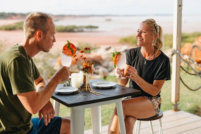 Two people sitting at an outdoor table with drinks and a vase of flowers. 2