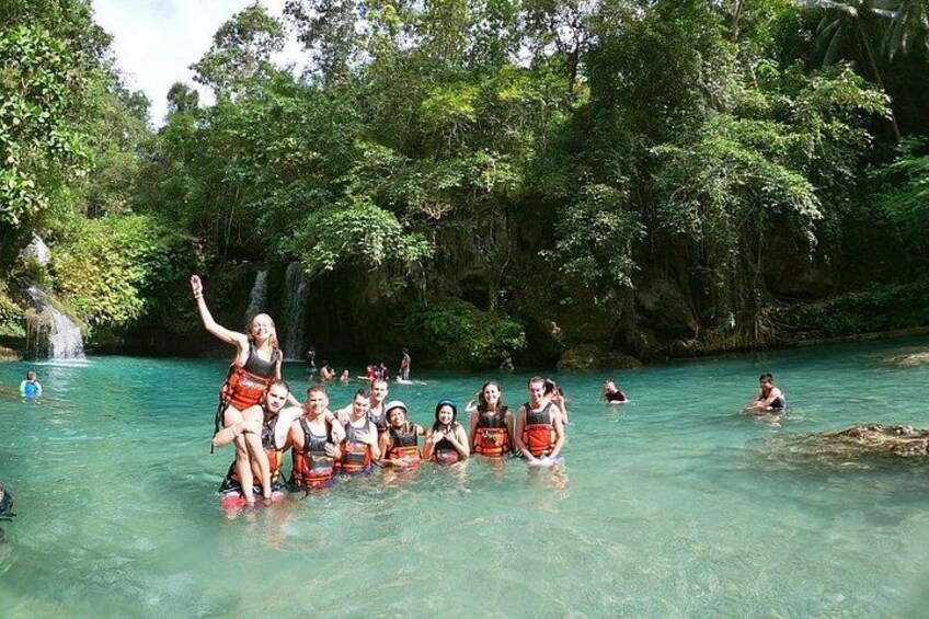 Een groep mensen in helder, turquoise water, omgeven door weelderige begroeiing en een waterval. 2