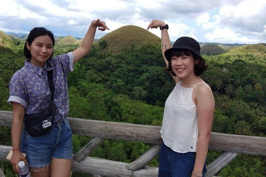 Two people standing on a wooden platform with a lush green landscape in the background. 1
