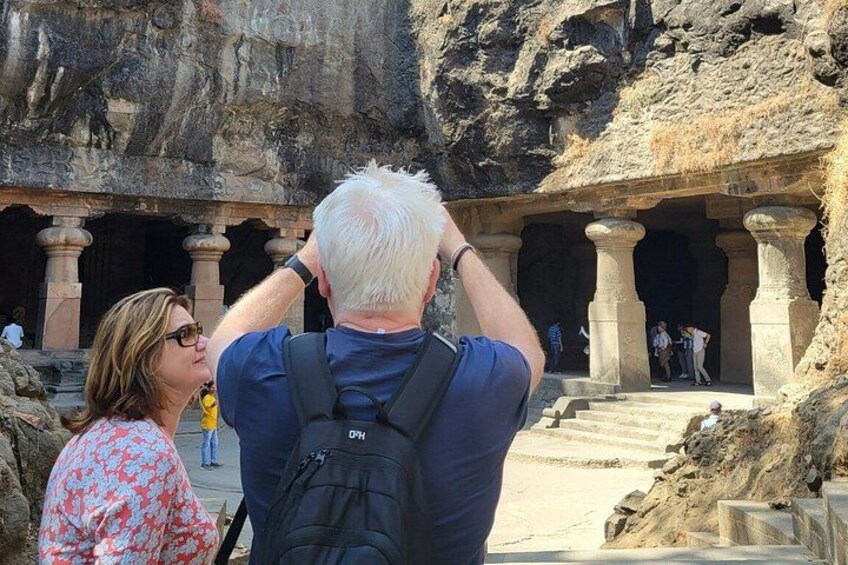 A man taking a photo with his back to the camera, standing in front of ancient rock-cut architecture with columns and steps. 4
