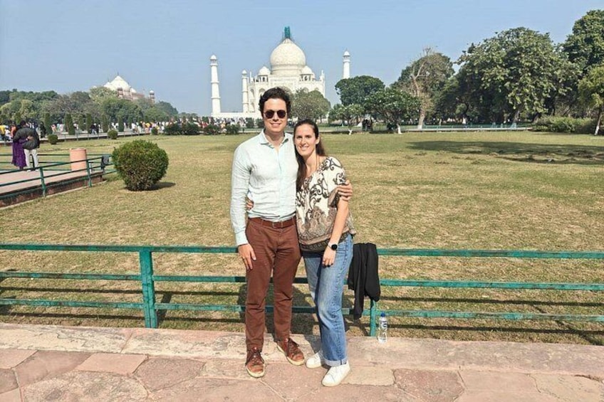 Una pareja de pie frente a un gran edificio blanco con cúpula y un minarete, rodeado por un parque con césped verde y árboles. 5
