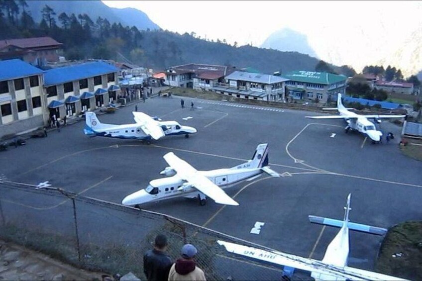 Una pista con varios aviones pequeños estacionados, un edificio con techo rojo y montañas al fondo. 1