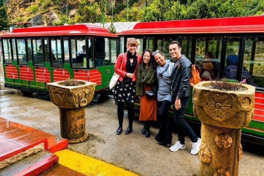 Un grupo de personas de pie frente a un tranvía rojo y verde, con dos grandes macetas de piedra en primer plano. 5