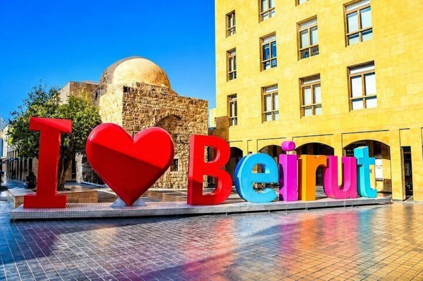 Een bord met 'I ♥ Beirut' in kleurrijke letters, een stenen gebouw en een helderblauwe lucht. 1