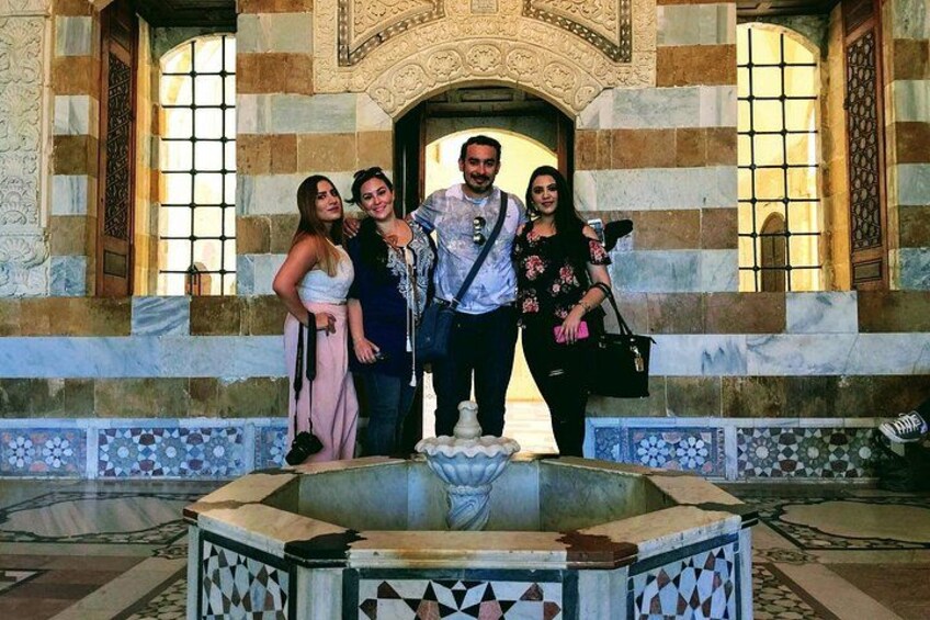 A group of people standing in front of a fountain with a tiled base and a building with arched windows and decorative stonework. 4