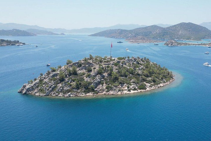 Eine Vogelperspektive einer kleinen Insel, umgeben von klarem, blauem Wasser, mit einer Flagge auf der Spitze und Booten in der Ferne. 1