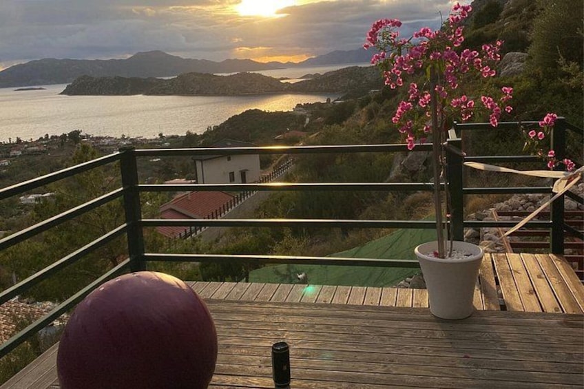 Eine Holzterrasse mit einem Blumentopf und einem roten Ball mit Blick auf eine Küstenlandschaft mit Hügeln und einem Gewässer im Sonnenuntergang. 4