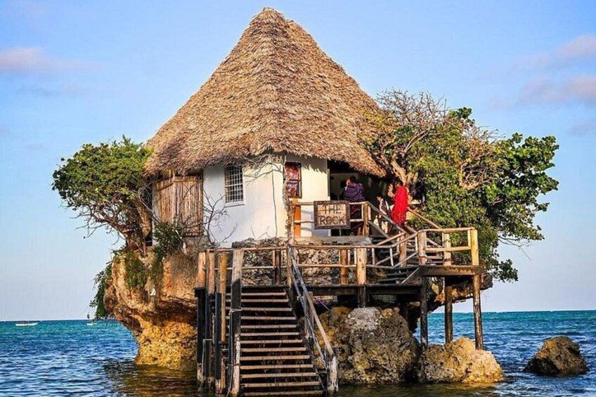 A thatched-roof house on a rocky outcrop with a wooden staircase leading up to it. 2