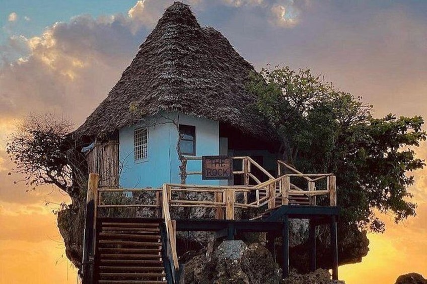 Una casa con tetto di paglia su palafitte nell'acqua, con un cartello che recita 'The Rock'. 1