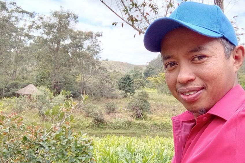 Une personne portant une chemise rose et une casquette bleue, debout dans un paysage naturel avec des arbres et un petit étang. 4