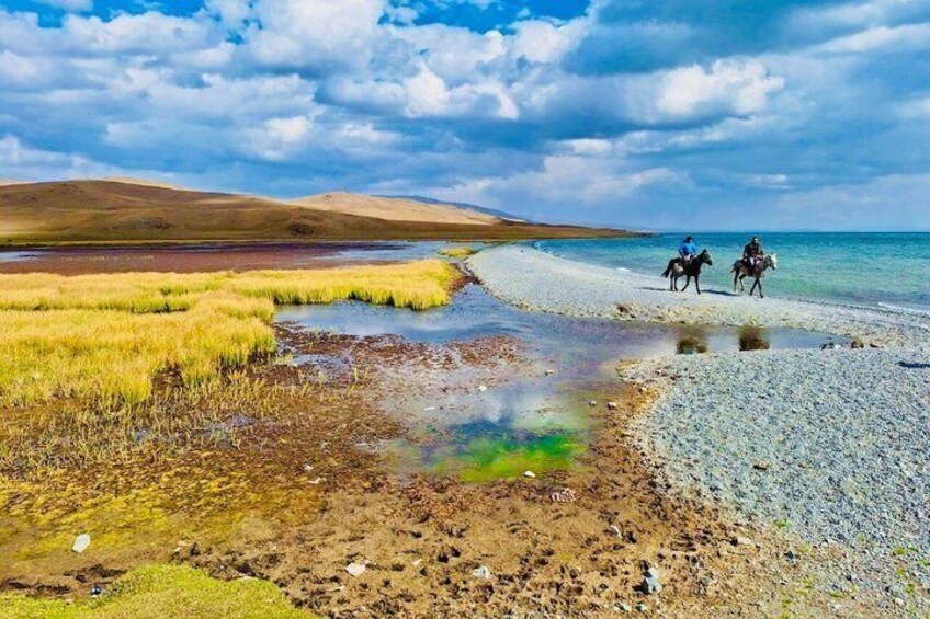 A riverbank with horses and riders, grassy areas, and distant hills. 2